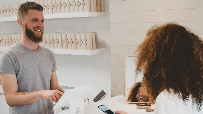 Come fidelizzare un cliente? - Uomo in maglietta grigia alla cassa di un negozio sorride a una cliente